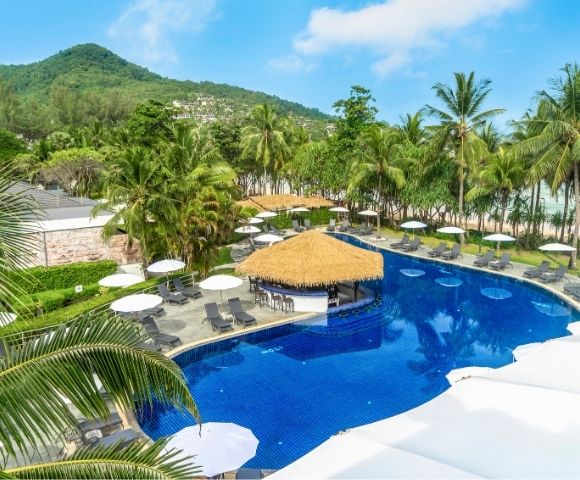 Resort pool surrounded by lounge chairs and umbrellas, with palm trees and a green mountain in the background under a clear blue sky.