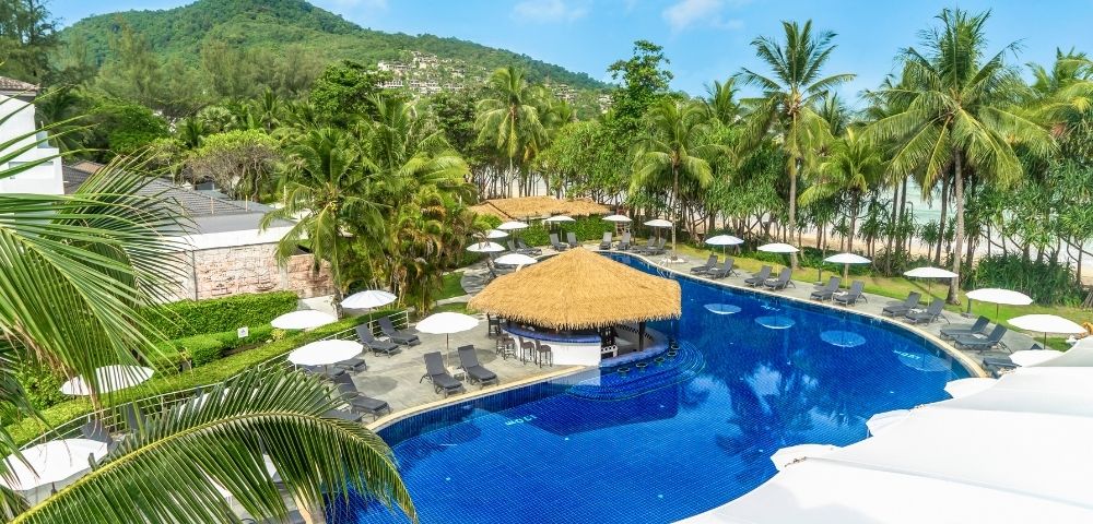 Resort pool surrounded by lounge chairs and umbrellas, with palm trees and a green mountain in the background under a clear blue sky.