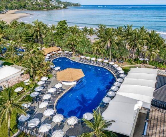 Aerial view of a luxurious beachfront resort with a curvy blue pool surrounded by white umbrellas and palm trees, adjacent to a serene sandy beach.