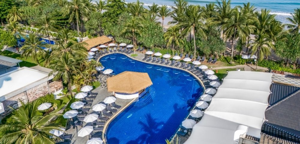 Aerial view of a luxurious beachfront resort with a curvy blue pool surrounded by white umbrellas and palm trees, adjacent to a serene sandy beach.