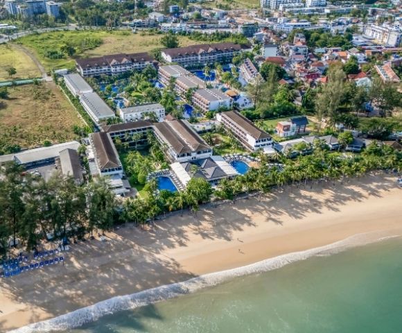 Aerial view of a beachfront resort with tropical trees, multiple pools, and sandy beach. The ocean meets the shore under a clear blue sky, creating a serene atmosphere.