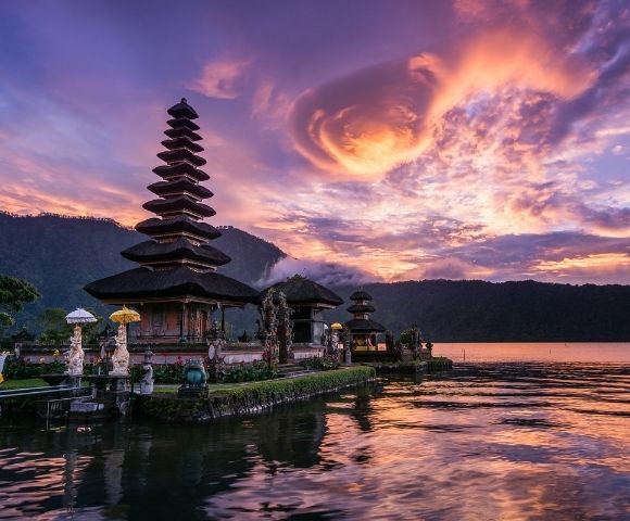 A multi-tiered Balinese temple complex with traditional architecture sits on the edge of a calm lake reflecting a vibrant sunset sky filled with dramatic clouds.