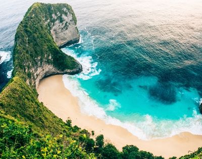 An aerial view of a dramatic green cliff overlooking a secluded sandy beach with turquoise water and crashing white waves.