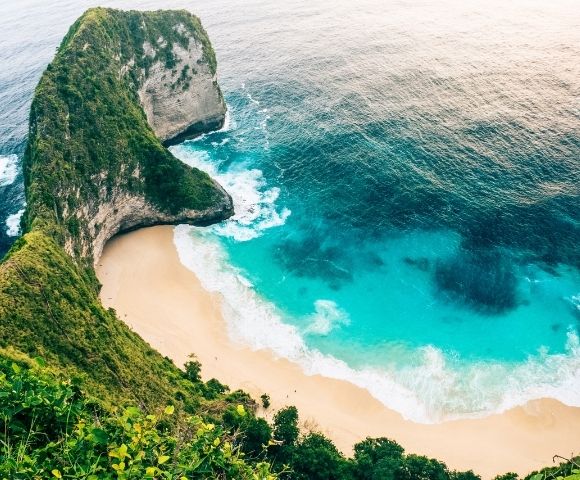 An aerial view of a dramatic green cliff overlooking a secluded sandy beach with turquoise water and crashing white waves.