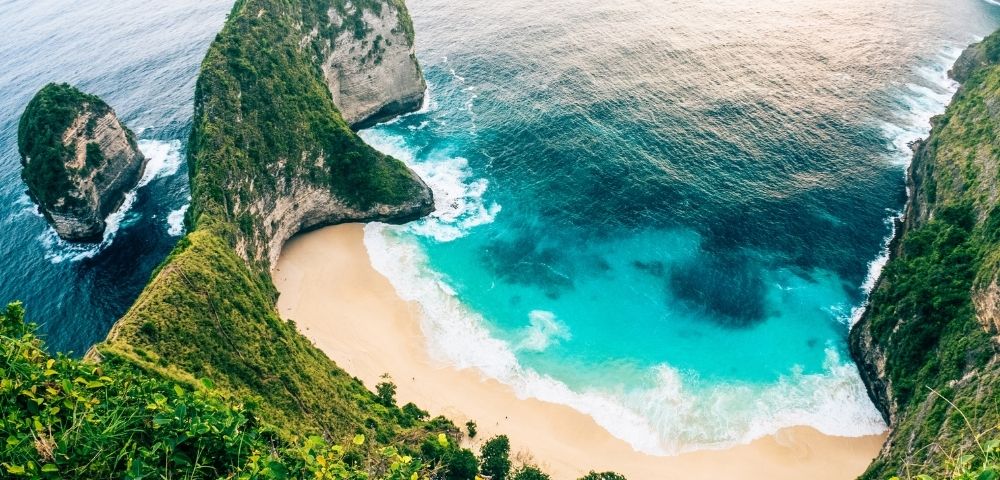 An aerial view of a dramatic green cliff overlooking a secluded sandy beach with turquoise water and crashing white waves.