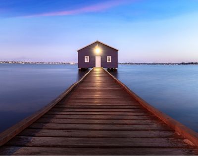 A purple boathouse sits at the end of a wooden pier over calm blue water at dusk, with a faint pink and blue twilight sky above.