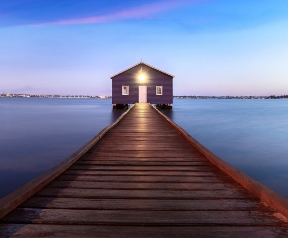 A purple boathouse sits at the end of a wooden pier over calm blue water at dusk, with a faint pink and blue twilight sky above.