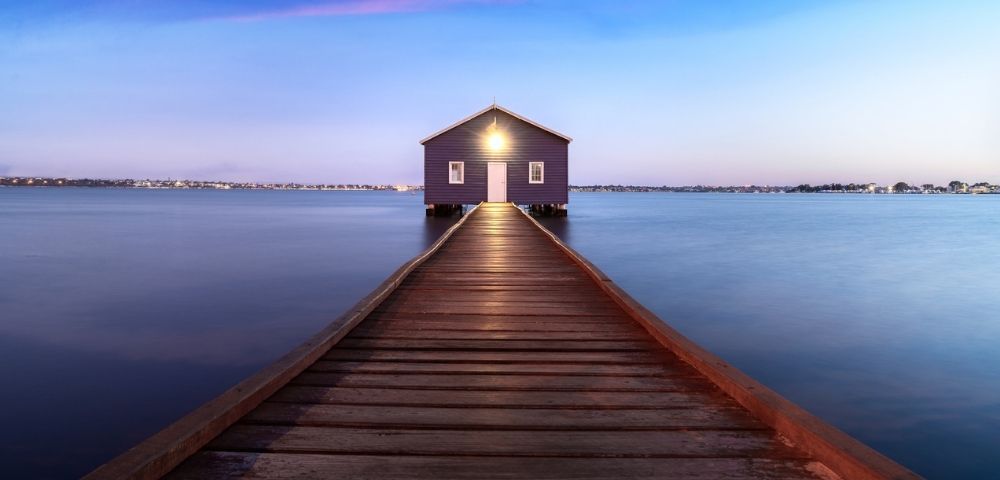 A purple boathouse sits at the end of a wooden pier over calm blue water at dusk, with a faint pink and blue twilight sky above.