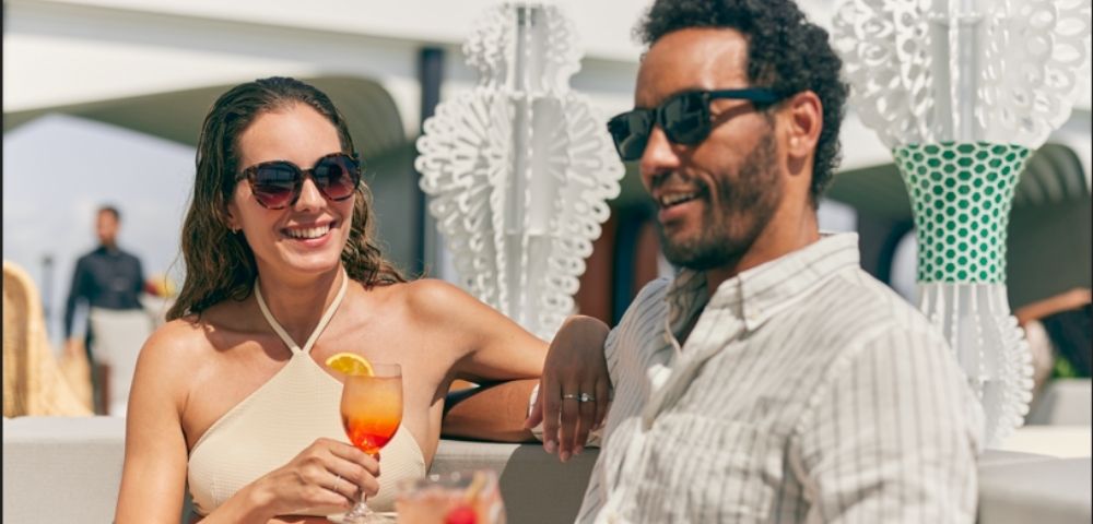 A man and a woman in sunglasses smile and converse at an outdoor social gathering, with a tropical drink in the foreground.