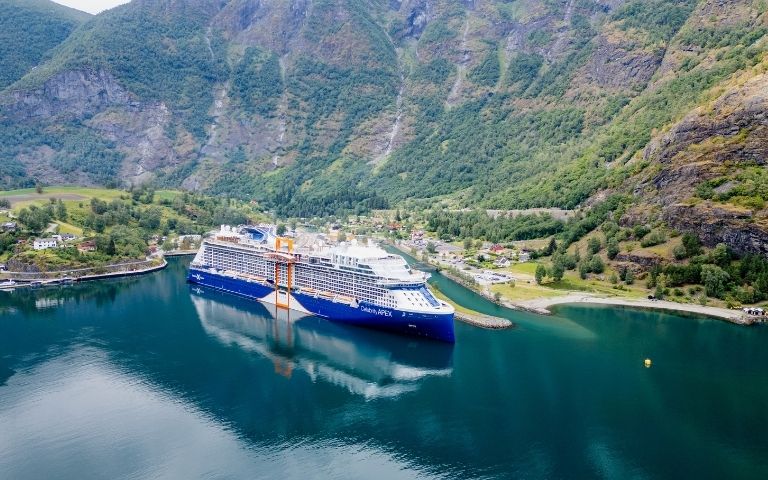A large blue cruise ship sails through a calm body of water, surrounded by steep, green mountains under a bright sky.