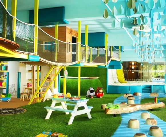 An indoor children's play area with a multi-level wooden and yellow structure, rope bridges, a yellow slide into a ball pit, and a green artificial turf floor.