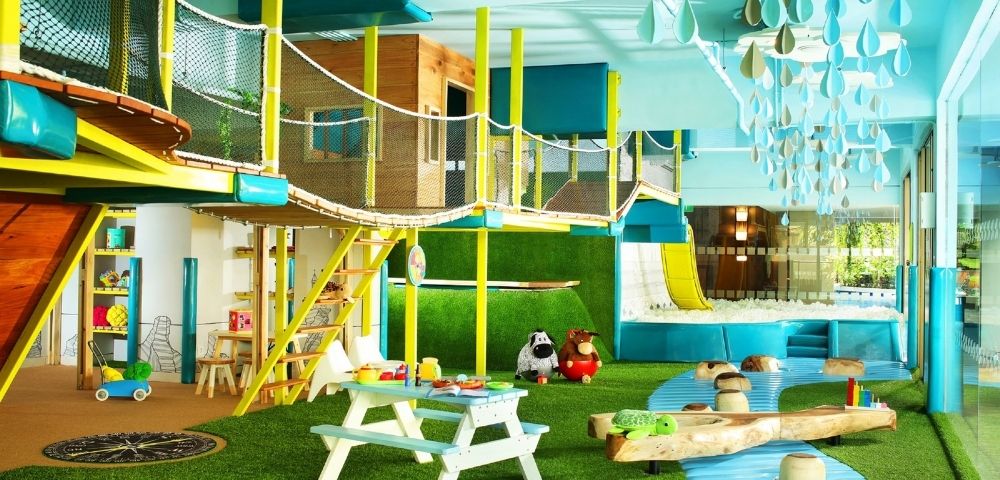An indoor children's play area with a multi-level wooden and yellow structure, rope bridges, a yellow slide into a ball pit, and a green artificial turf floor.