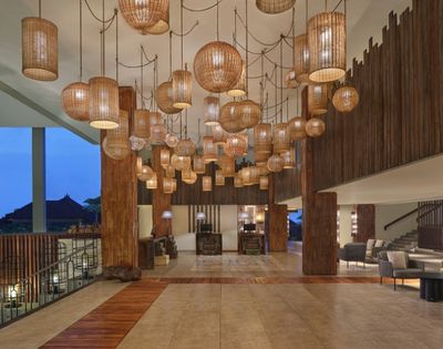 A hotel lobby featuring numerous rattan pendant lights of various shapes and sizes hanging from the ceiling, with wood-paneled walls, a tiled floor with wood accents, and a reception area in the background.