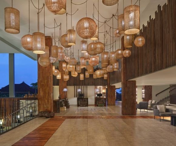 A hotel lobby featuring numerous rattan pendant lights of various shapes and sizes hanging from the ceiling, with wood-paneled walls, a tiled floor with wood accents, and a reception area in the background.