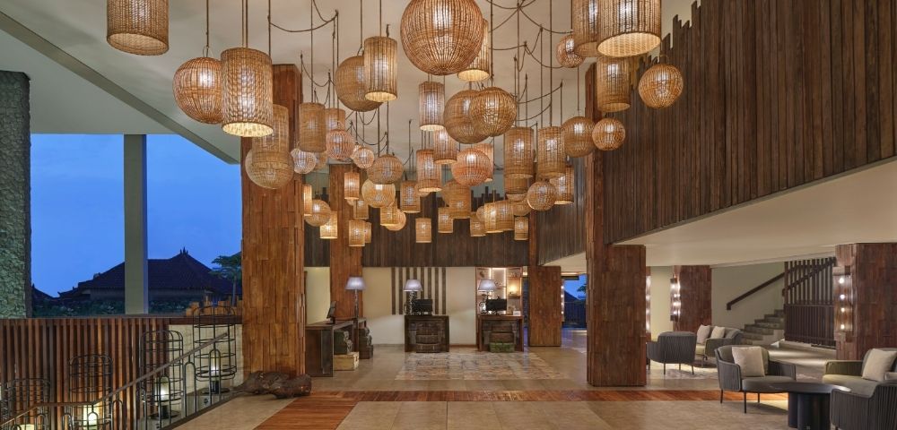 A hotel lobby featuring numerous rattan pendant lights of various shapes and sizes hanging from the ceiling, with wood-paneled walls, a tiled floor with wood accents, and a reception area in the background.