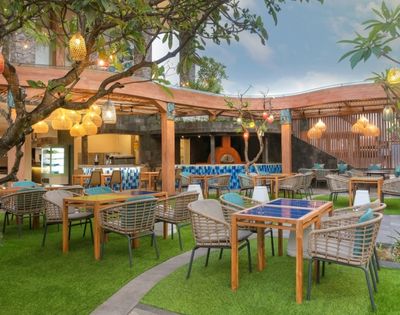 An outdoor restaurant seating area with tables and chairs arranged on artificial turf, with a wooden pergola overhead, decorative lanterns hanging from trees, and a pizza oven visible in the background.
