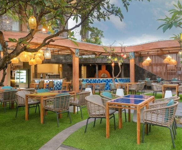 An outdoor restaurant seating area with tables and chairs arranged on artificial turf, with a wooden pergola overhead, decorative lanterns hanging from trees, and a pizza oven visible in the background.