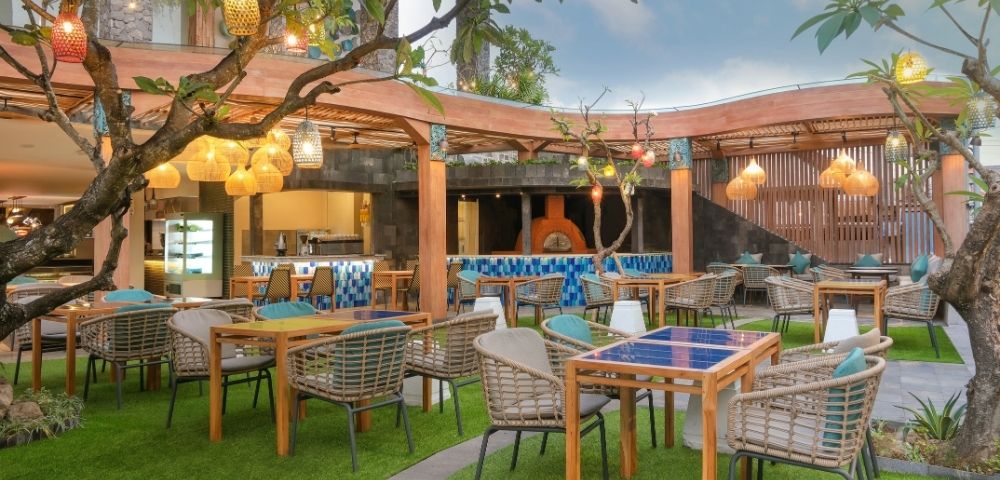 An outdoor restaurant seating area with tables and chairs arranged on artificial turf, with a wooden pergola overhead, decorative lanterns hanging from trees, and a pizza oven visible in the background.