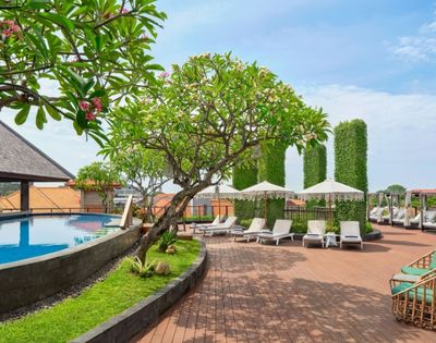 A tropical resort's rooftop pool area with lounge chairs, umbrellas, and lush greenery under a bright blue sky.