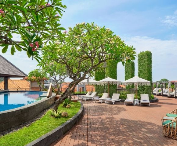 A tropical resort's rooftop pool area with lounge chairs, umbrellas, and lush greenery under a bright blue sky.