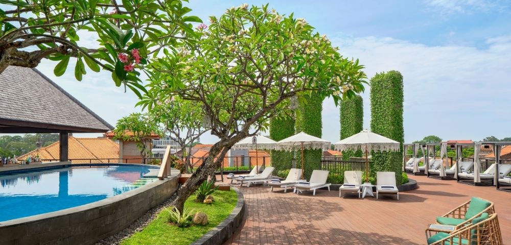 A tropical resort's rooftop pool area with lounge chairs, umbrellas, and lush greenery under a bright blue sky.