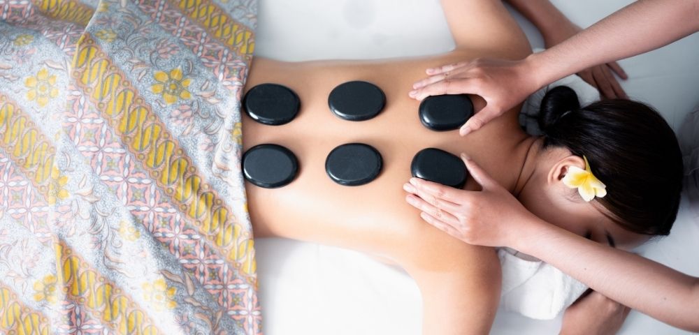 Overhead view of a person receiving a hot stone massage on their back, with dark smooth stones arranged in a grid pattern and a floral patterned cloth draped over their legs.