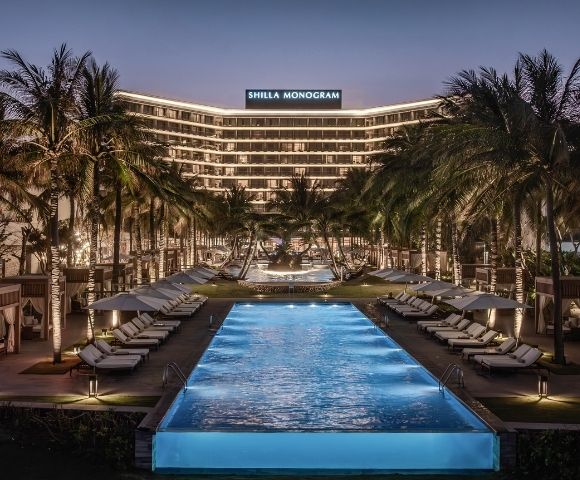 A luxury resort at dusk with a rectangular swimming pool in the foreground, rows of lounge chairs with umbrellas, private cabanas, and palm trees lining the path leading to a modern, illuminated hotel building with 