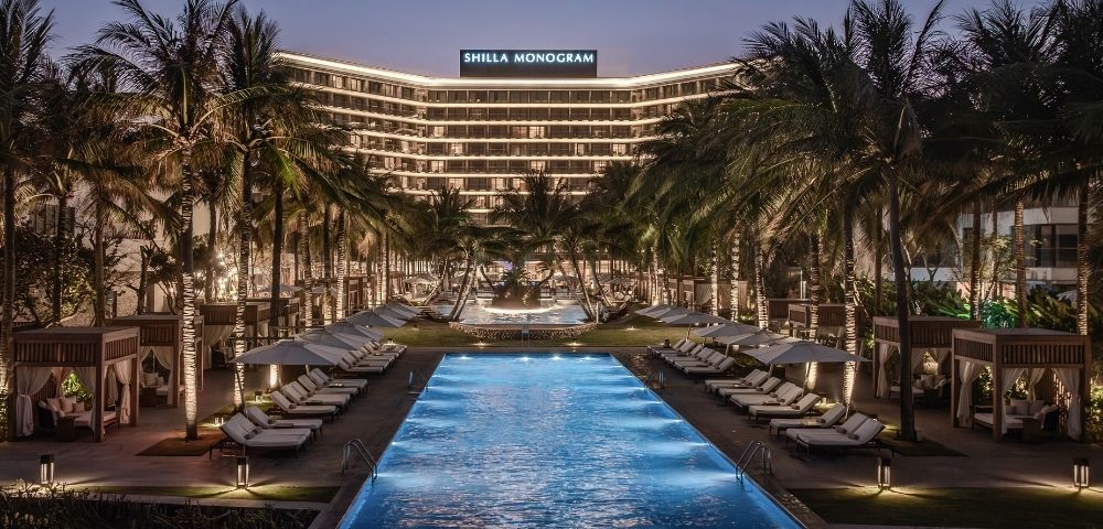 A luxury resort at dusk with a rectangular swimming pool in the foreground, rows of lounge chairs with umbrellas, private cabanas, and palm trees lining the path leading to a modern, illuminated hotel building with 