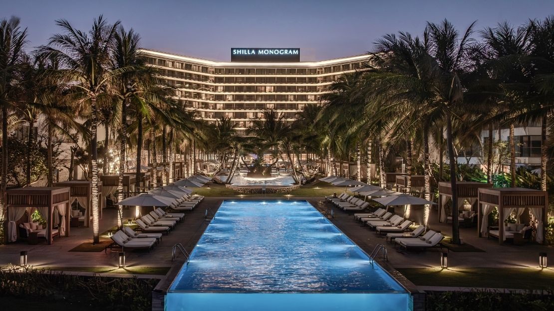 A luxury resort at dusk with a rectangular swimming pool in the foreground, rows of lounge chairs with umbrellas, private cabanas, and palm trees lining the path leading to a modern, illuminated hotel building with 