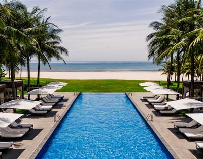 An infinity swimming pool at a tropical resort, lined with lounge chairs and umbrellas, overlooks a sandy beach and the ocean.