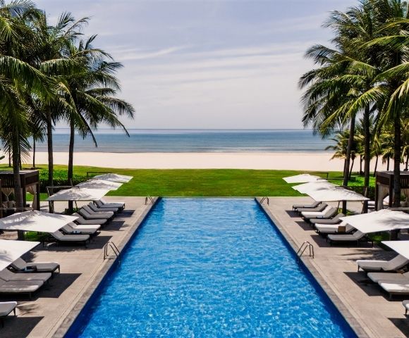 An infinity swimming pool at a tropical resort, lined with lounge chairs and umbrellas, overlooks a sandy beach and the ocean.