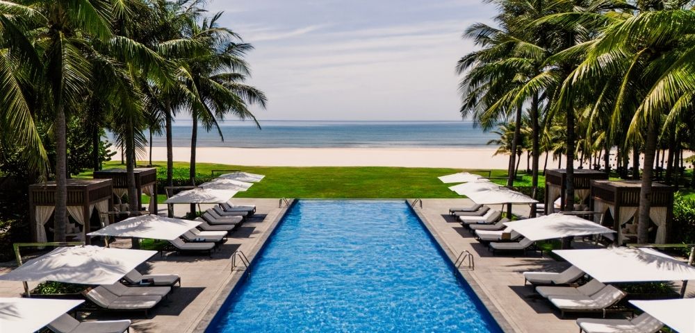 An infinity swimming pool at a tropical resort, lined with lounge chairs and umbrellas, overlooks a sandy beach and the ocean.