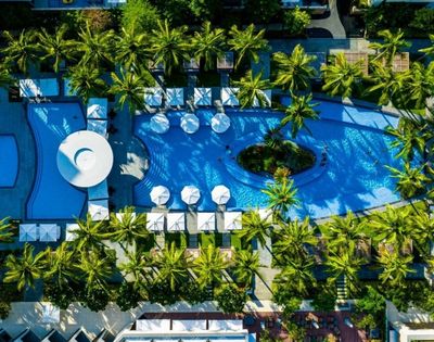 Aerial view of a resort complex featuring a large, curved swimming pool with lounge chairs and umbrellas surrounding it, amidst lush palm trees and manicured green spaces.
