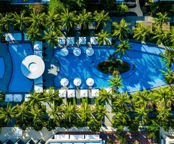 Aerial view of a resort complex featuring a large, curved swimming pool with lounge chairs and umbrellas surrounding it, amidst lush palm trees and manicured green spaces.