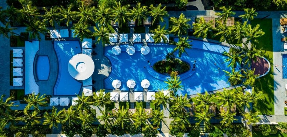 Aerial view of a resort complex featuring a large, curved swimming pool with lounge chairs and umbrellas surrounding it, amidst lush palm trees and manicured green spaces.
