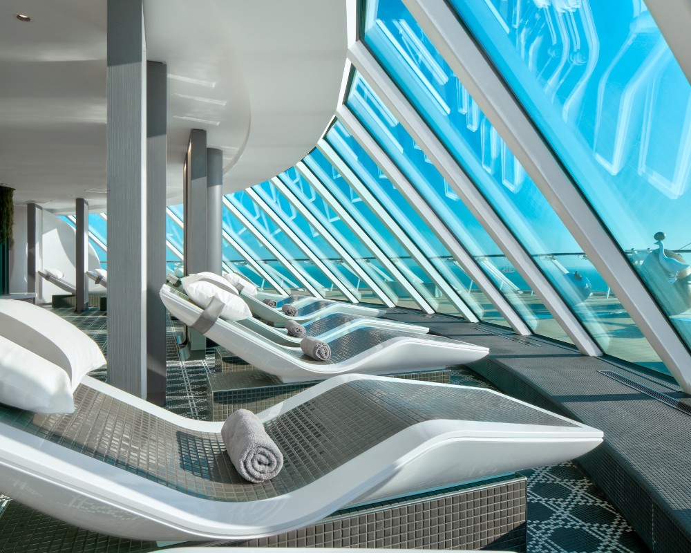 Row of modern lounge chairs with rolled towels and pillows by large angled windows, in a bright, serene relaxation area.
