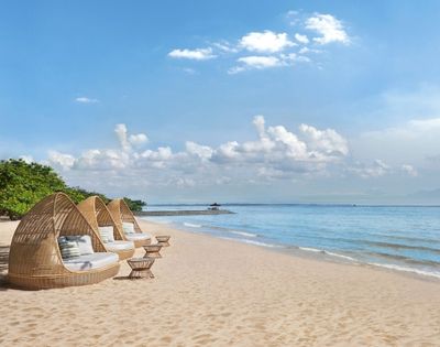 Three woven daybeds with cushions and small woven tables sit on a sandy beach facing a calm blue ocean under a sky with white clouds.