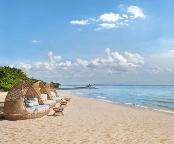 Three woven daybeds with cushions and small woven tables sit on a sandy beach facing a calm blue ocean under a sky with white clouds.