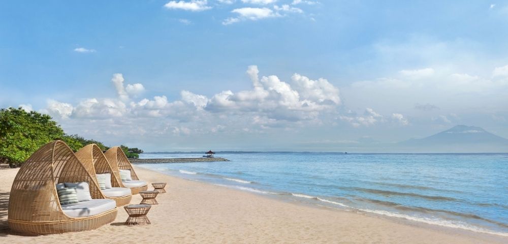 Three woven daybeds with cushions and small woven tables sit on a sandy beach facing a calm blue ocean under a sky with white clouds.