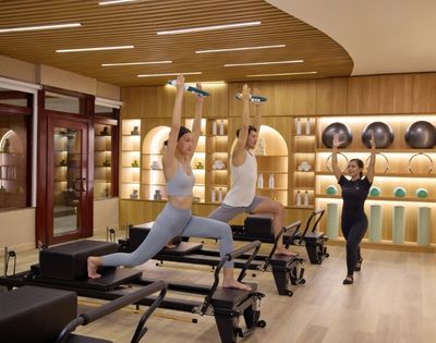 Three individuals practice Pilates on reformers in a well-lit studio with wooden accents and shelves of exercise equipment.