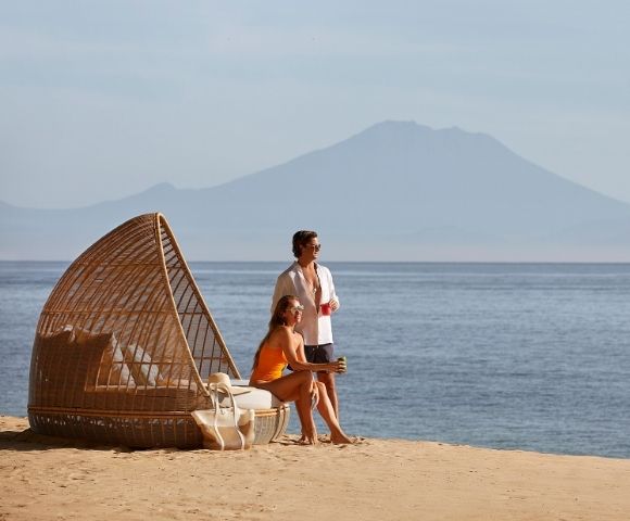 A couple relaxes on a sandy beach next to a woven outdoor daybed, with the calm ocean and a hazy mountain range in the background.