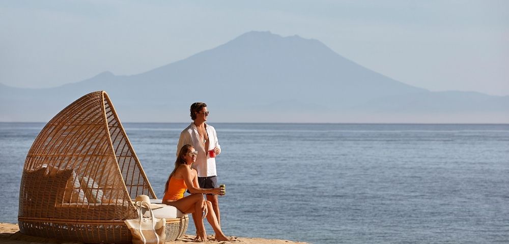 A couple relaxes on a sandy beach next to a woven outdoor daybed, with the calm ocean and a hazy mountain range in the background.