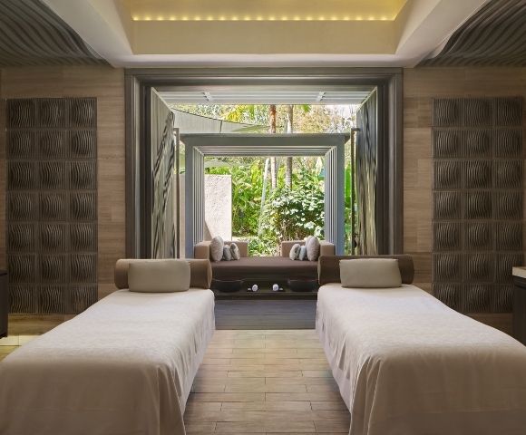 Two massage tables with light-colored linens in a spa room, with open doors revealing a lush garden view.