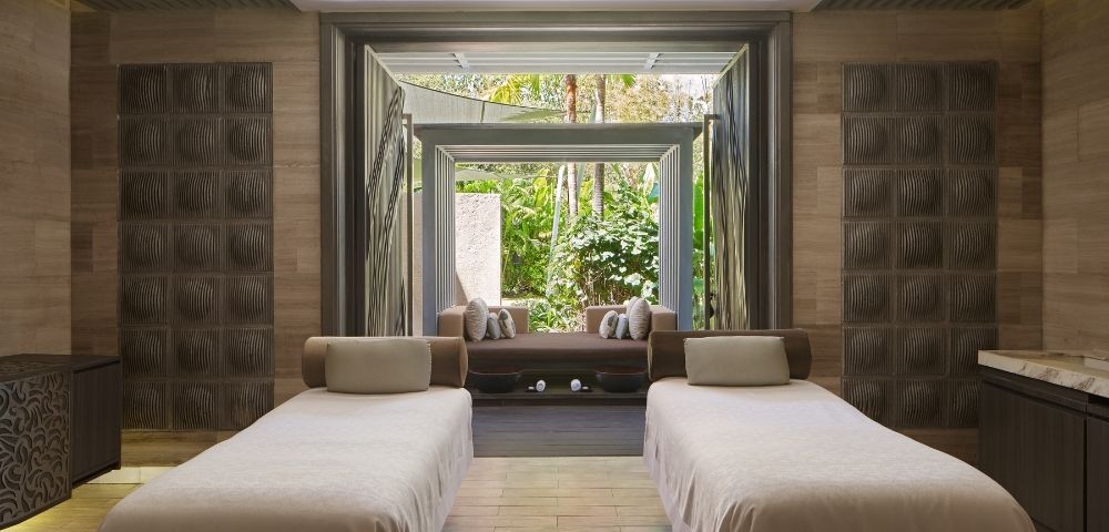 Two massage tables with light-colored linens in a spa room, with open doors revealing a lush garden view.