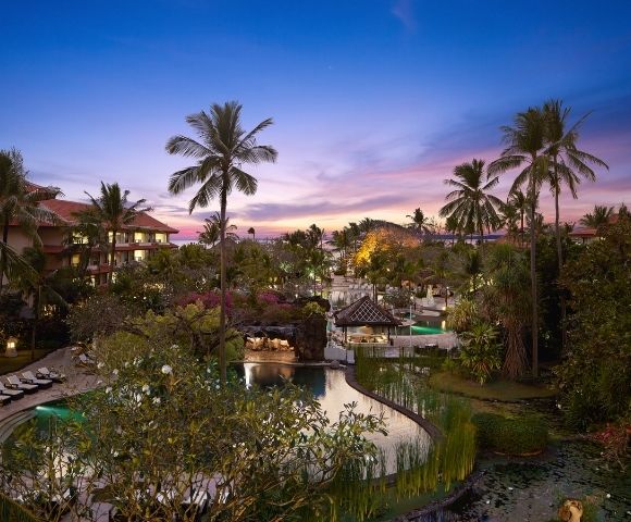 Tropical resort with multiple pools, palm trees, and lush greenery at dusk under a colorful sky.