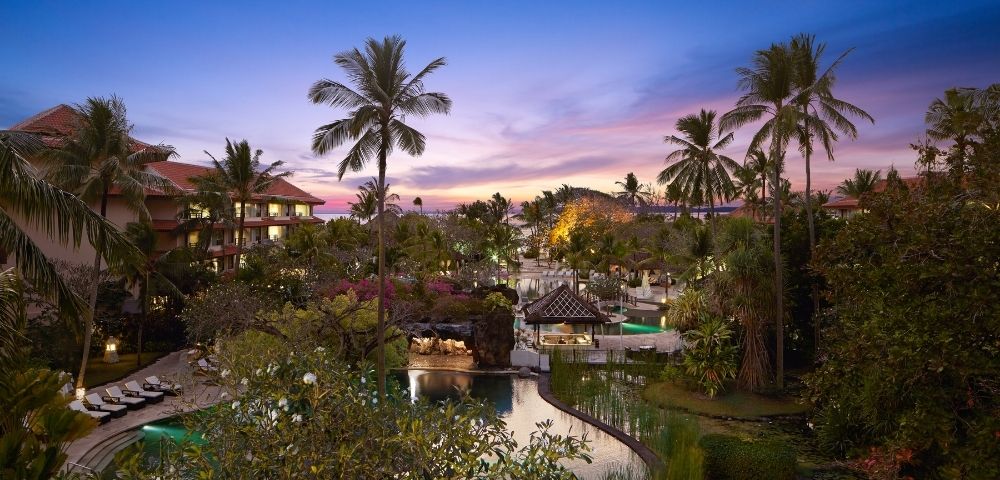 Tropical resort with multiple pools, palm trees, and lush greenery at dusk under a colorful sky.
