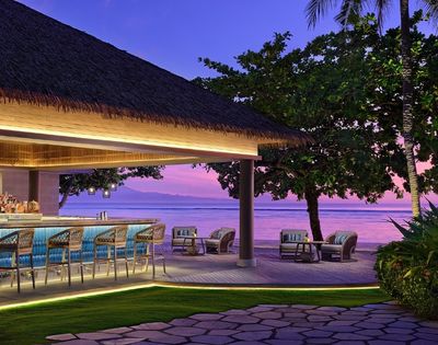 Outdoor tropical beach bar with wicker stools and lounge chairs at sunset, overlooking calm ocean and distant mountains.