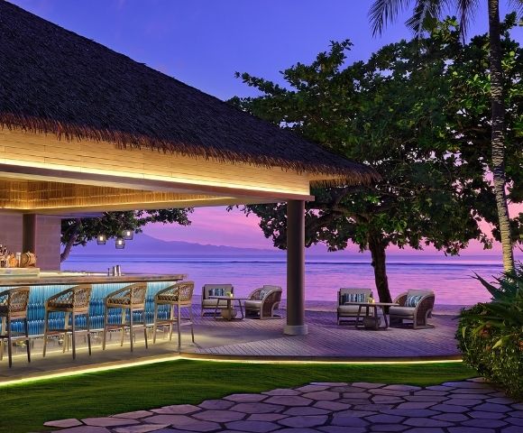 Outdoor tropical beach bar with wicker stools and lounge chairs at sunset, overlooking calm ocean and distant mountains.