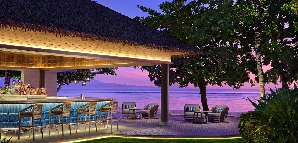 Outdoor tropical beach bar with wicker stools and lounge chairs at sunset, overlooking calm ocean and distant mountains.