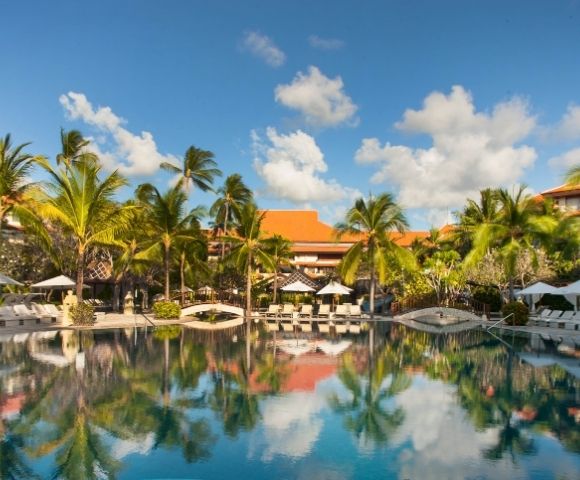 Resort swimming pool surrounded by palm trees, lounge chairs, umbrellas, and red-roofed buildings under a blue sky with scattered clouds.